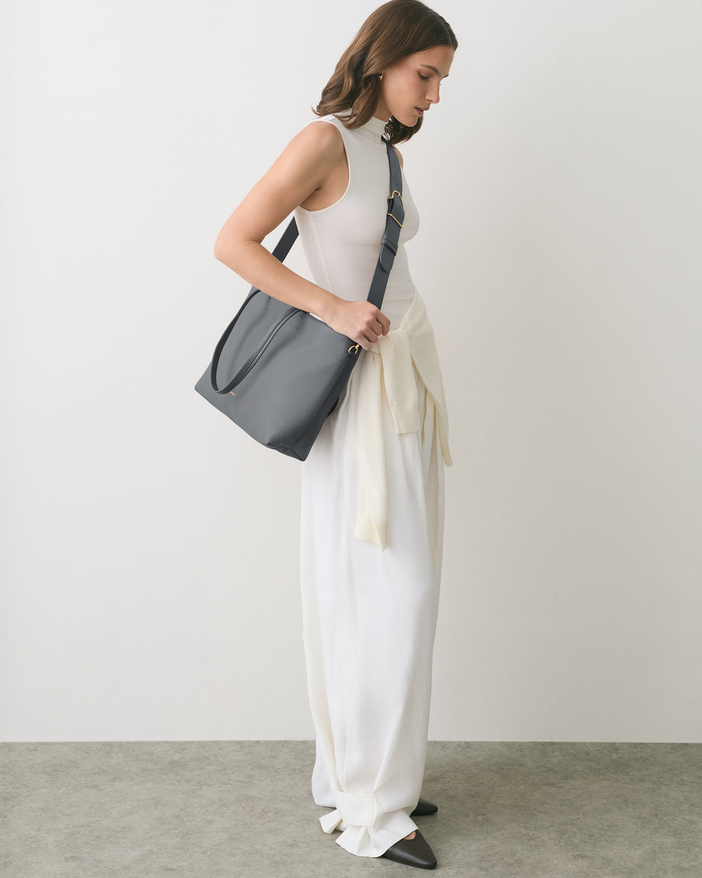 Woman standing with travel bag over shoulder, wearing long dress, sweater tied at waist
