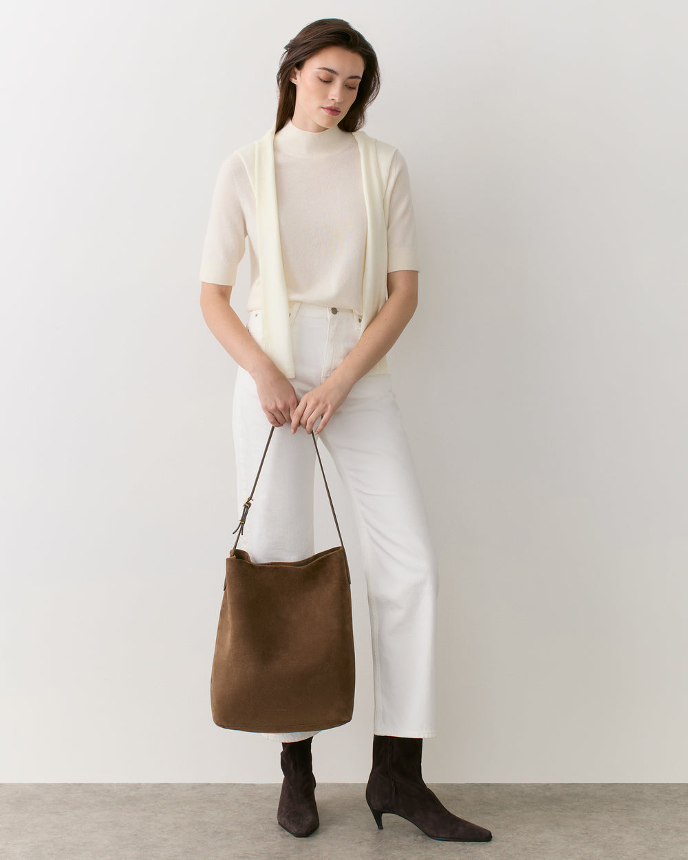 Person standing with a shoulder bag wearing a light top and pants, hands holding the bag strap