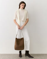 Person standing with a shoulder bag wearing a light top and pants, hands holding the bag strap