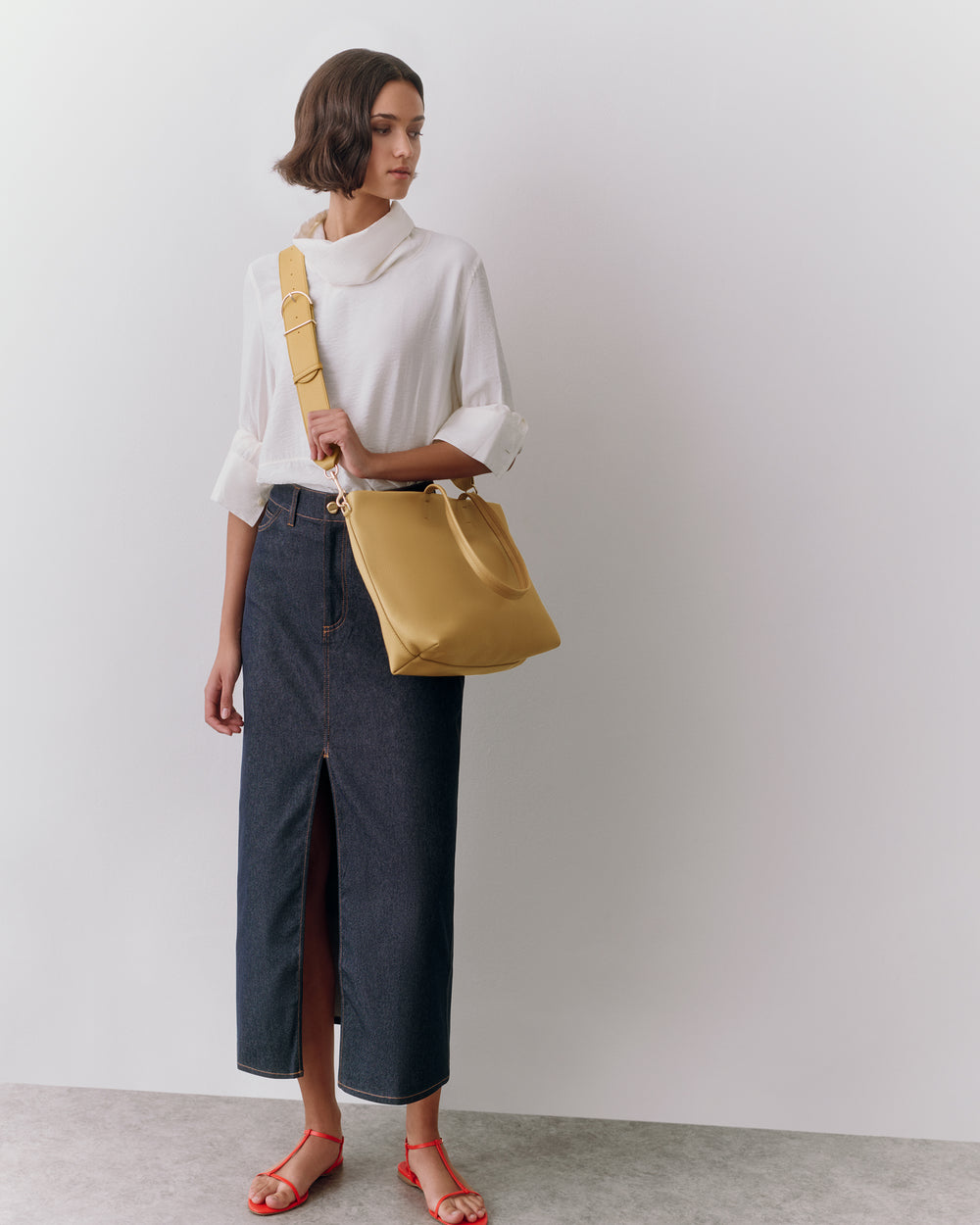 Woman in long skirt and top holding a shoulder bag standing against plain background.