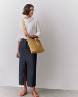 Woman in long skirt and top holding a shoulder bag standing against plain background.