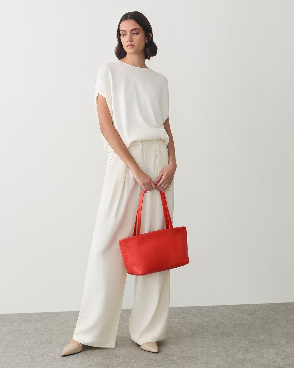 Woman in flowing outfit holding a tote bag standing on a plain floor surface.