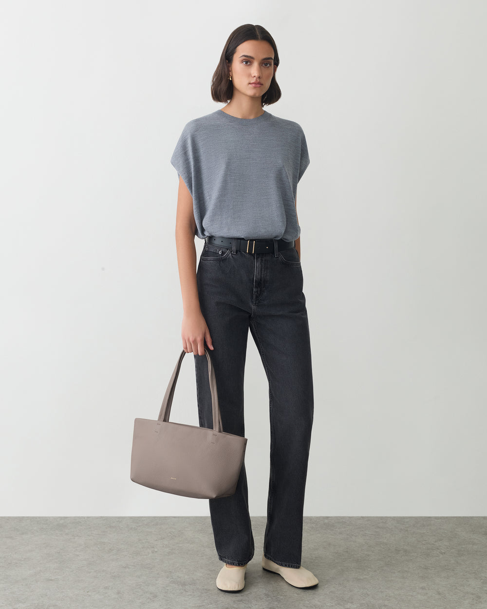 Person in a shirt and jeans holding a handbag standing on a plain background.