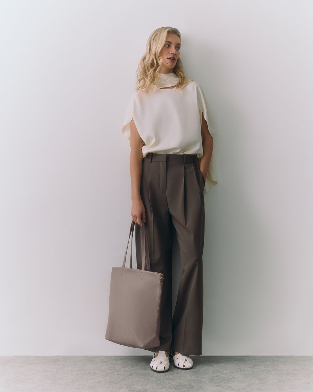 Person standing against wall holding tote bag wearing wide-leg pants and sleeveless top.