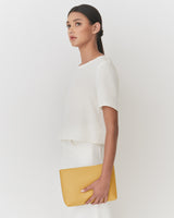 Person holding a clutch under one arm, wearing a short-sleeve top and looking to the side.