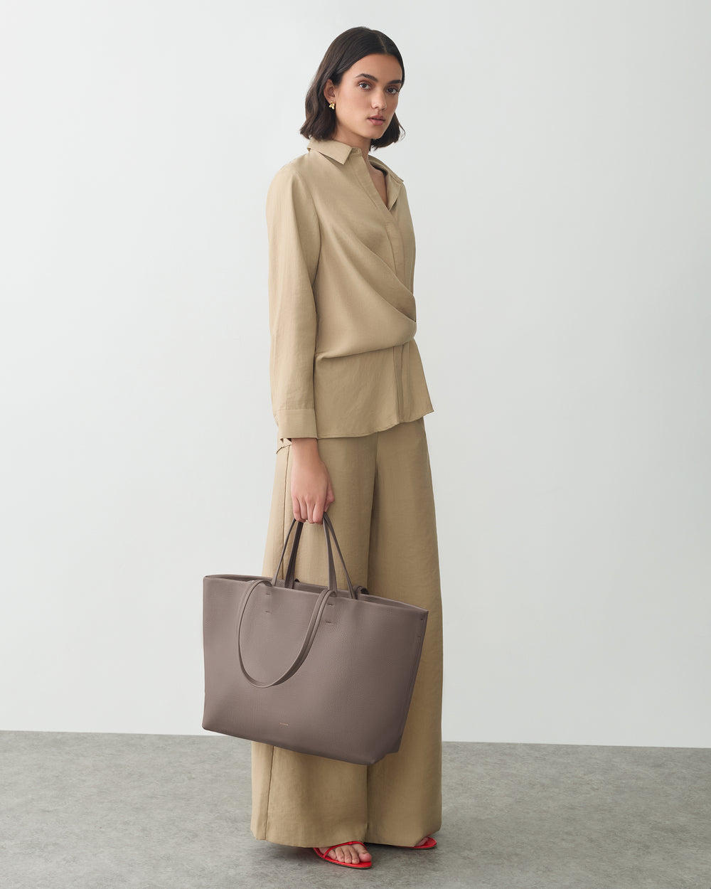 Person standing with travel bag, wearing relaxed outfit, against plain background.