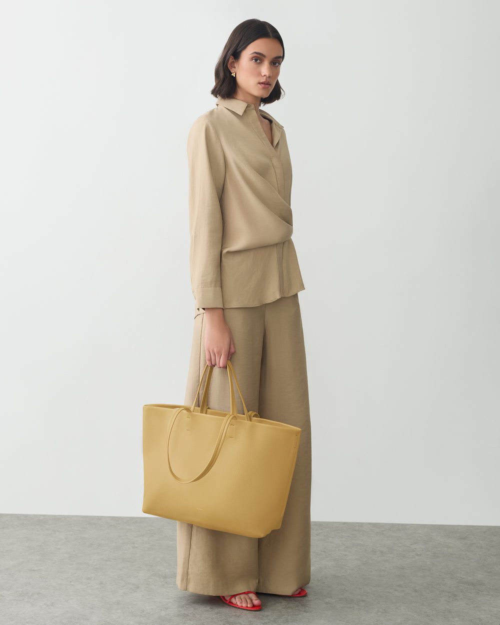Person in relaxed outfit holding a large handbag standing indoors.