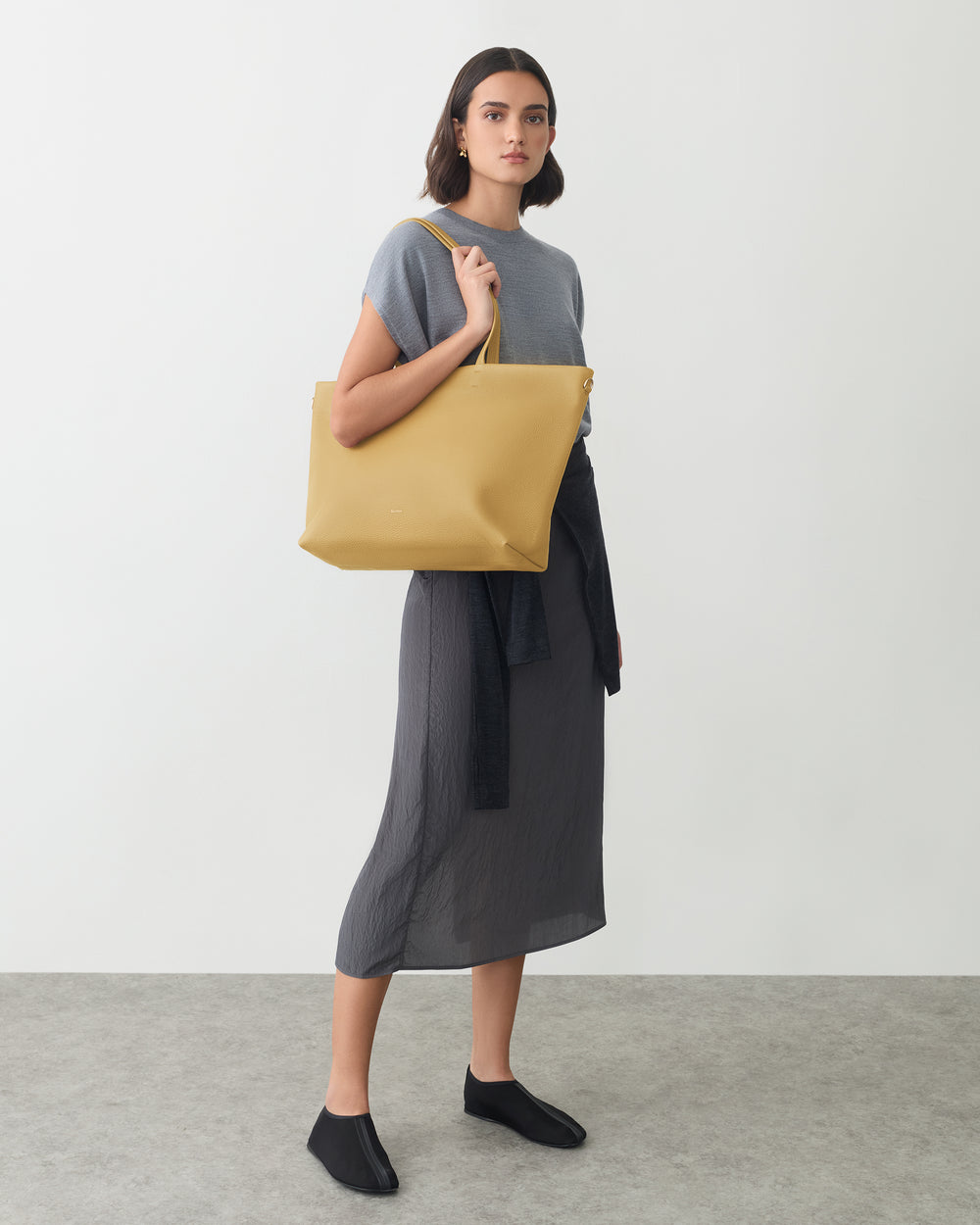 Person wearing a dress, holding a large tote bag, sweater tied at the waist.