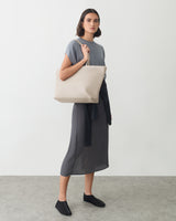 A person stands holding a large tote bag, wearing a shirt and skirt with a sweater wrapped around their waist.