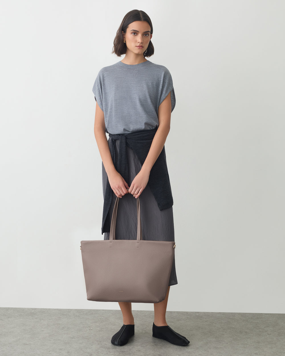 Person holding a handbag, wearing a short-sleeve top, skirt, and a sweater tied at the waist.
