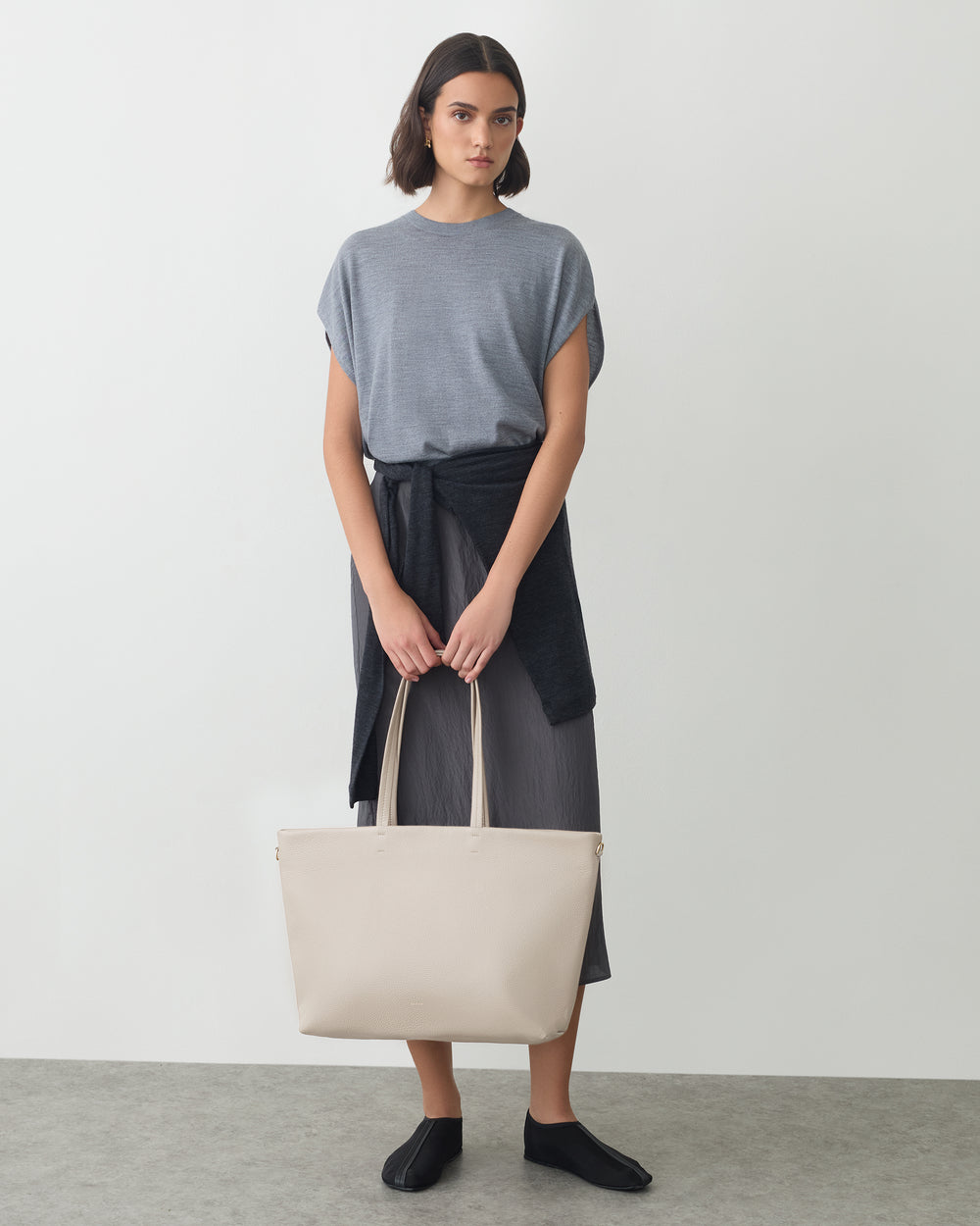 Person standing with large tote bag, shirt tied at waist, wearing a simple outfit and shoes.