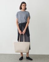 Person standing with large tote bag, shirt tied at waist, wearing a simple outfit and shoes.