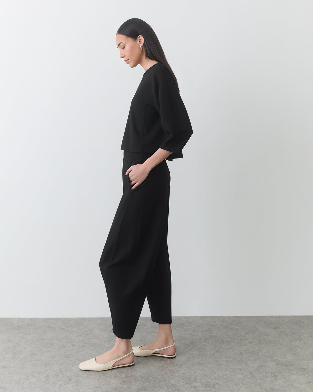 Person wearing a two-piece outfit and flat shoes, standing side profile in a plain room.