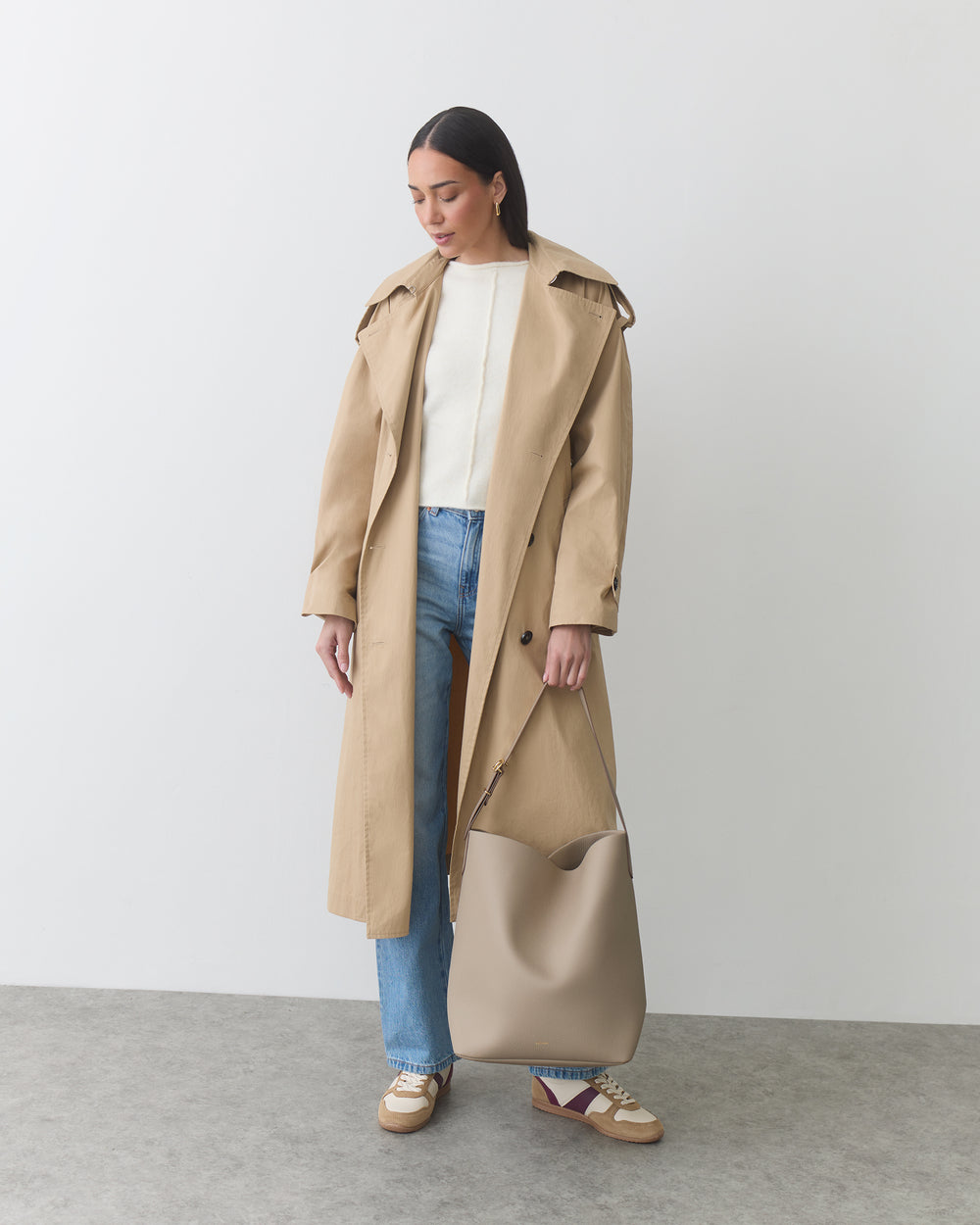 Woman in trench coat holding shoulder bag by its strap against a plain background.