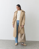 Woman in trench coat holding shoulder bag by its strap against a plain background.