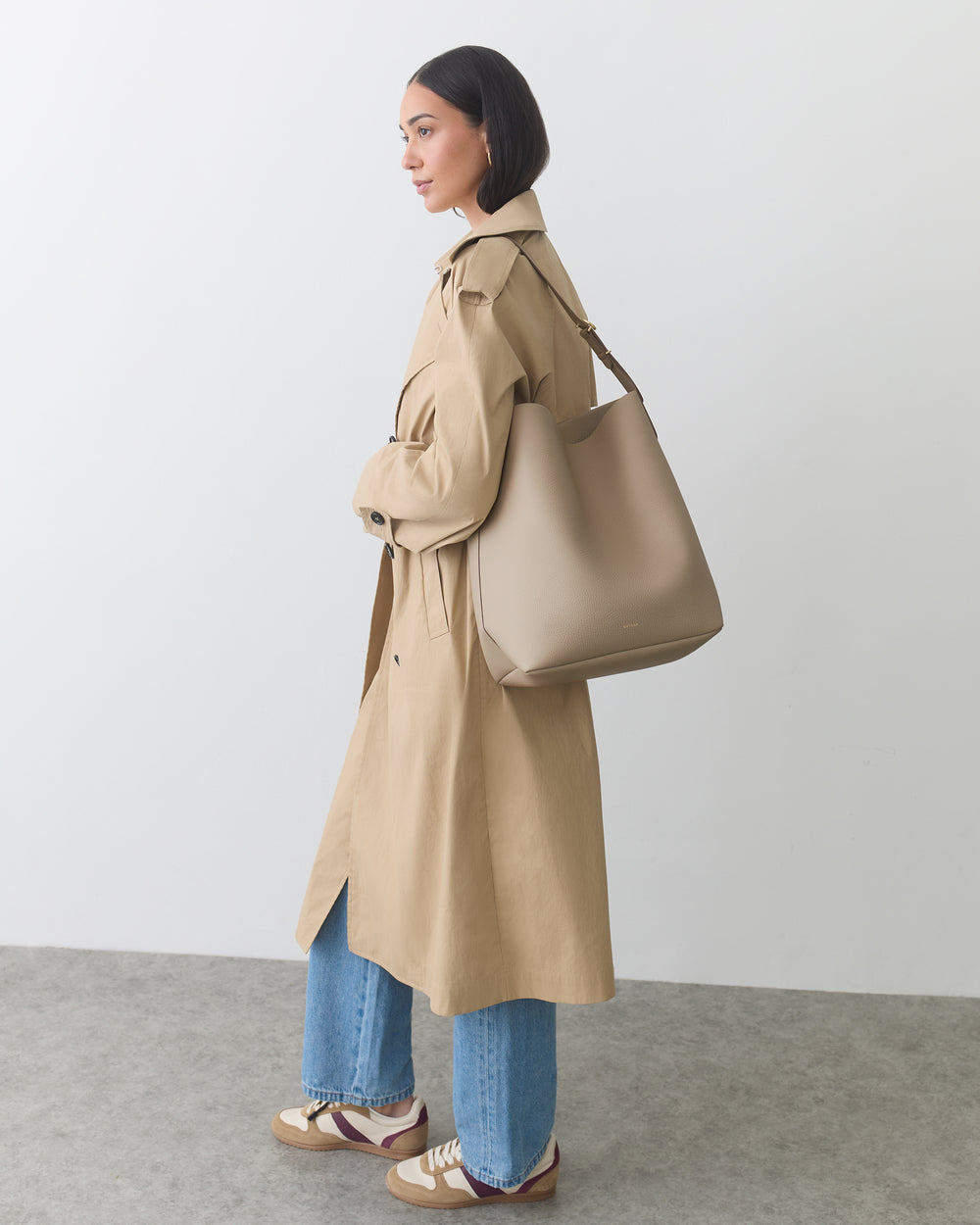 Woman in trench coat carrying a shoulder bag against a plain background.