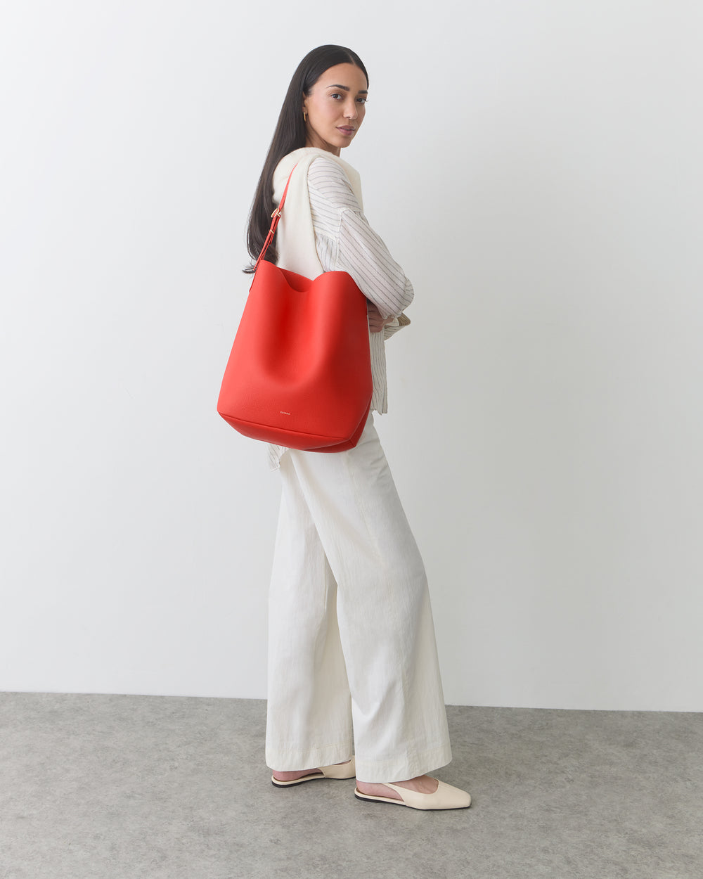 Woman standing in a neutral outfit carrying a brightly colored shoulder bag against a plain background.