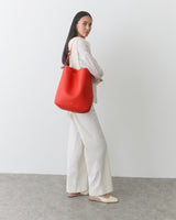 Woman standing in a neutral outfit carrying a brightly colored shoulder bag against a plain background.