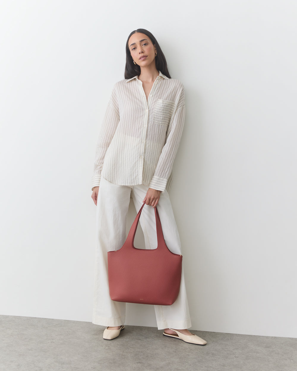 A person stands holding a large handbag, wearing a long-sleeve shirt and wide-leg pants.