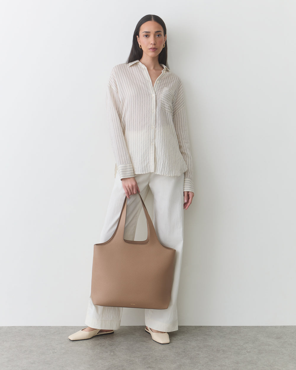 A person stands holding a large handbag, wearing a shirt and wide pants.