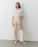 Woman standing against a wall holding a small handbag, wearing a shirt and wide-leg pants.