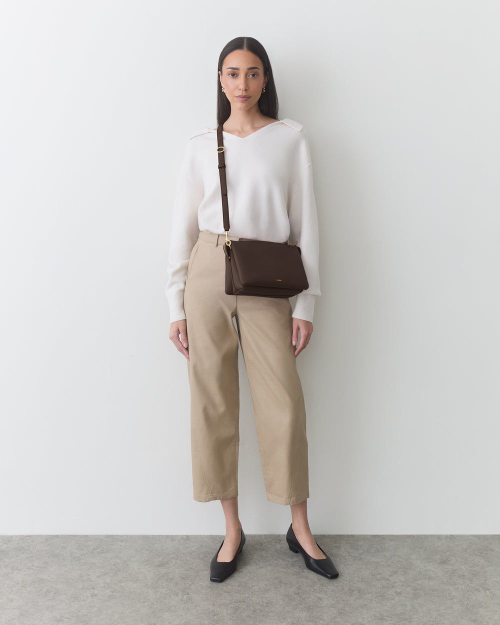 Person in sweater and pants with a crossbody bag, standing against a plain background.