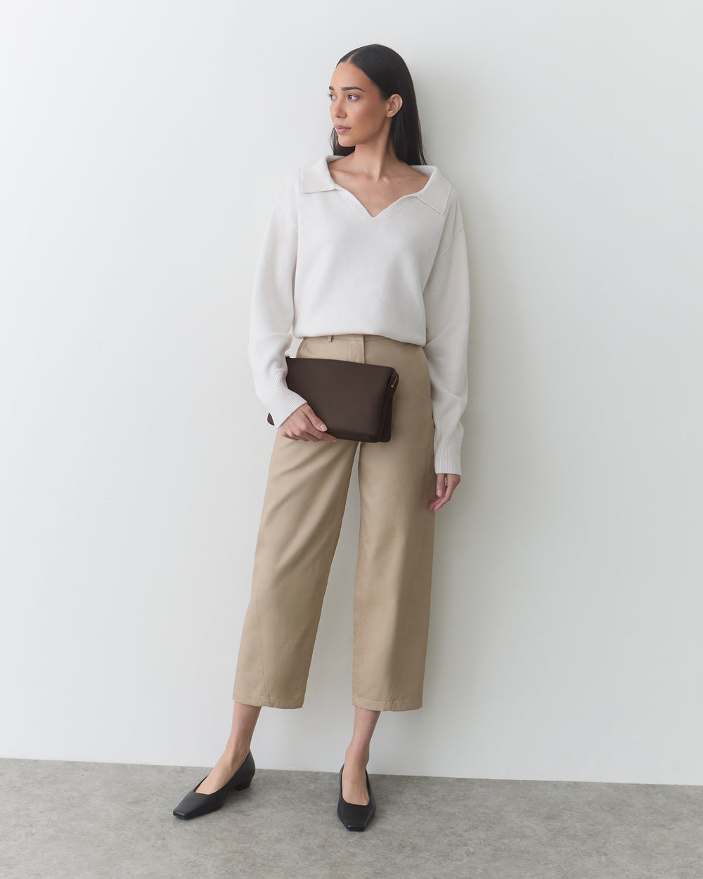 Person standing with long sleeves, cropped pants, holding a clutch bag.