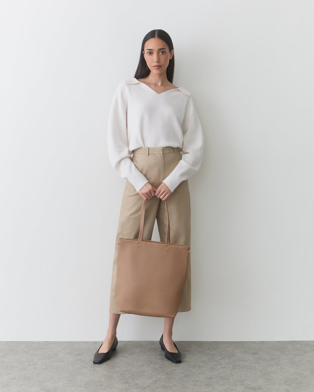 Woman standing with large tote bag, wearing sweater and wide-leg pants.
