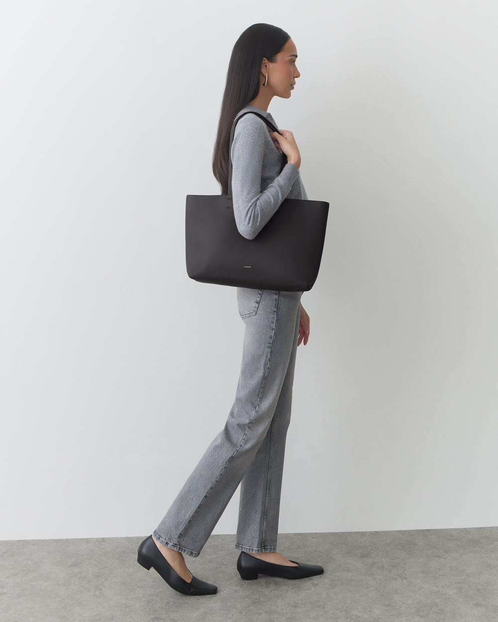 Person standing with a tote bag on shoulder, wearing a long-sleeve top and pants.