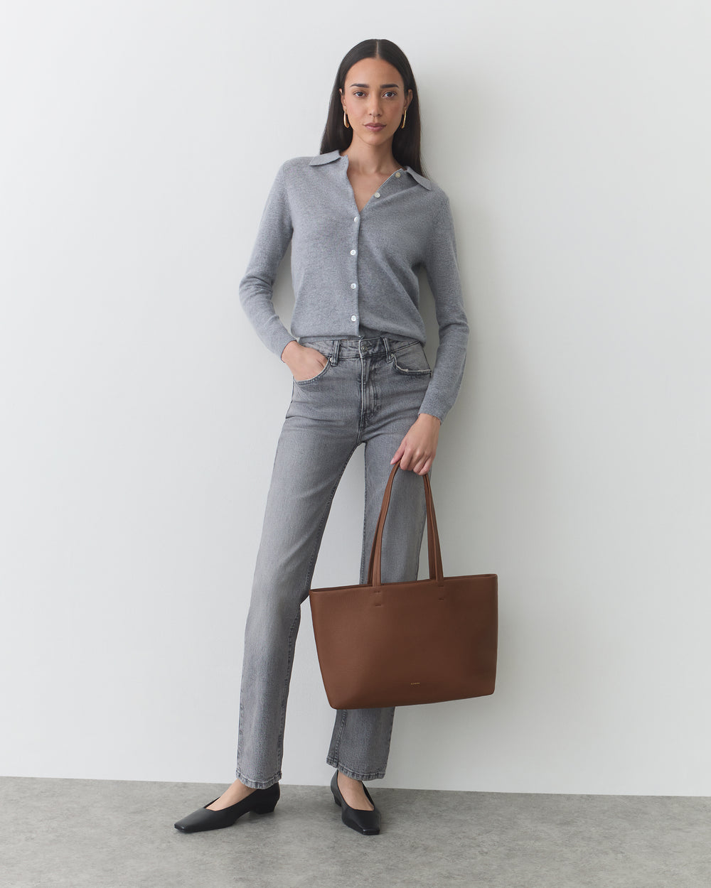 Woman standing, wearing button-up shirt, jeans, flats, holding a shoulder bag.