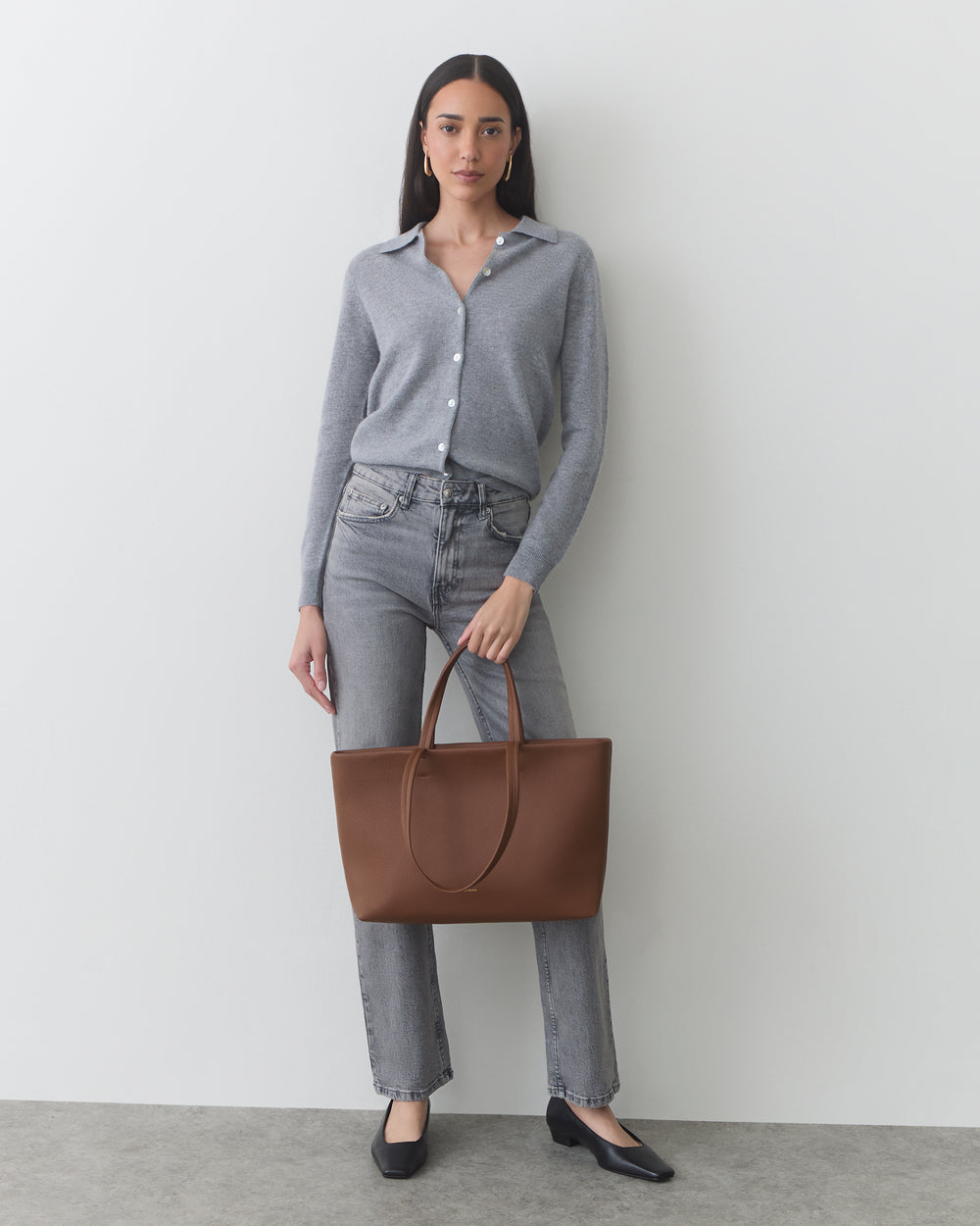 Woman in a shirt and jeans holding a tote bag, standing against a plain backdrop.