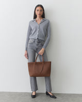 Woman in a shirt and jeans holding a tote bag, standing against a plain backdrop.
