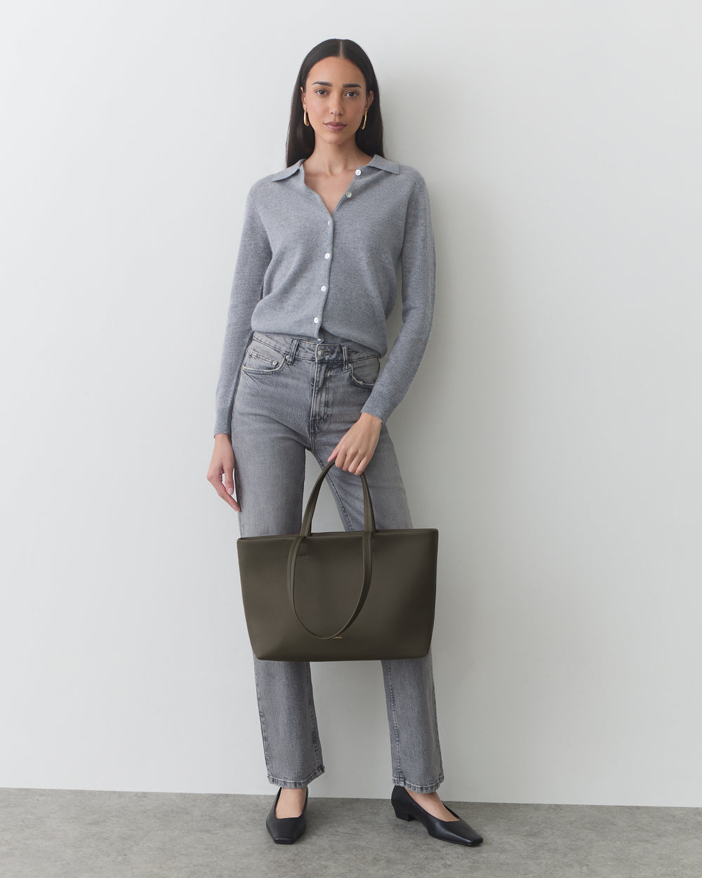 Person standing against a wall holding a large handbag, wearing a long-sleeve top and jeans.