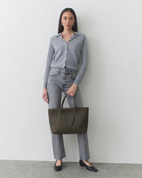 Person standing against a wall holding a large handbag, wearing a long-sleeve top and jeans.