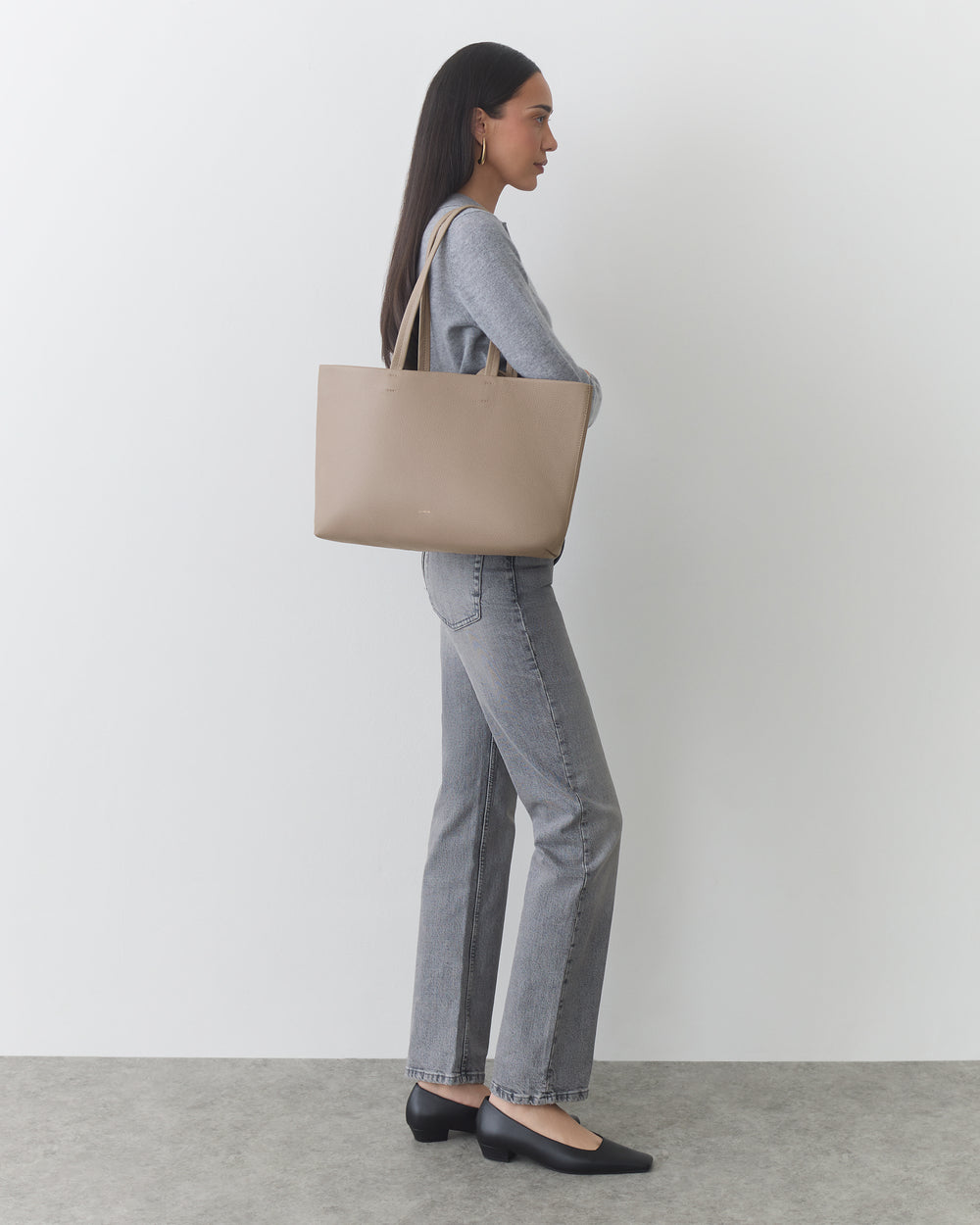 Person with long hair holding a tote bag, wearing a top, jeans, and shoes, standing sideways.
