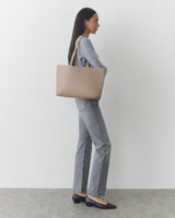 Person with long hair holding a tote bag, wearing a top, jeans, and shoes, standing sideways.