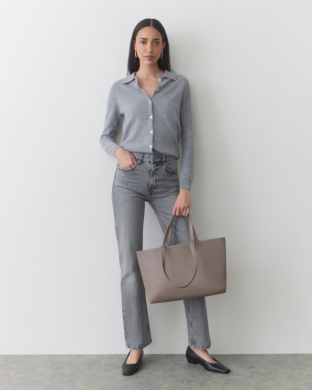 Woman in button-up shirt and jeans holding a large handbag, standing against a plain backdrop.