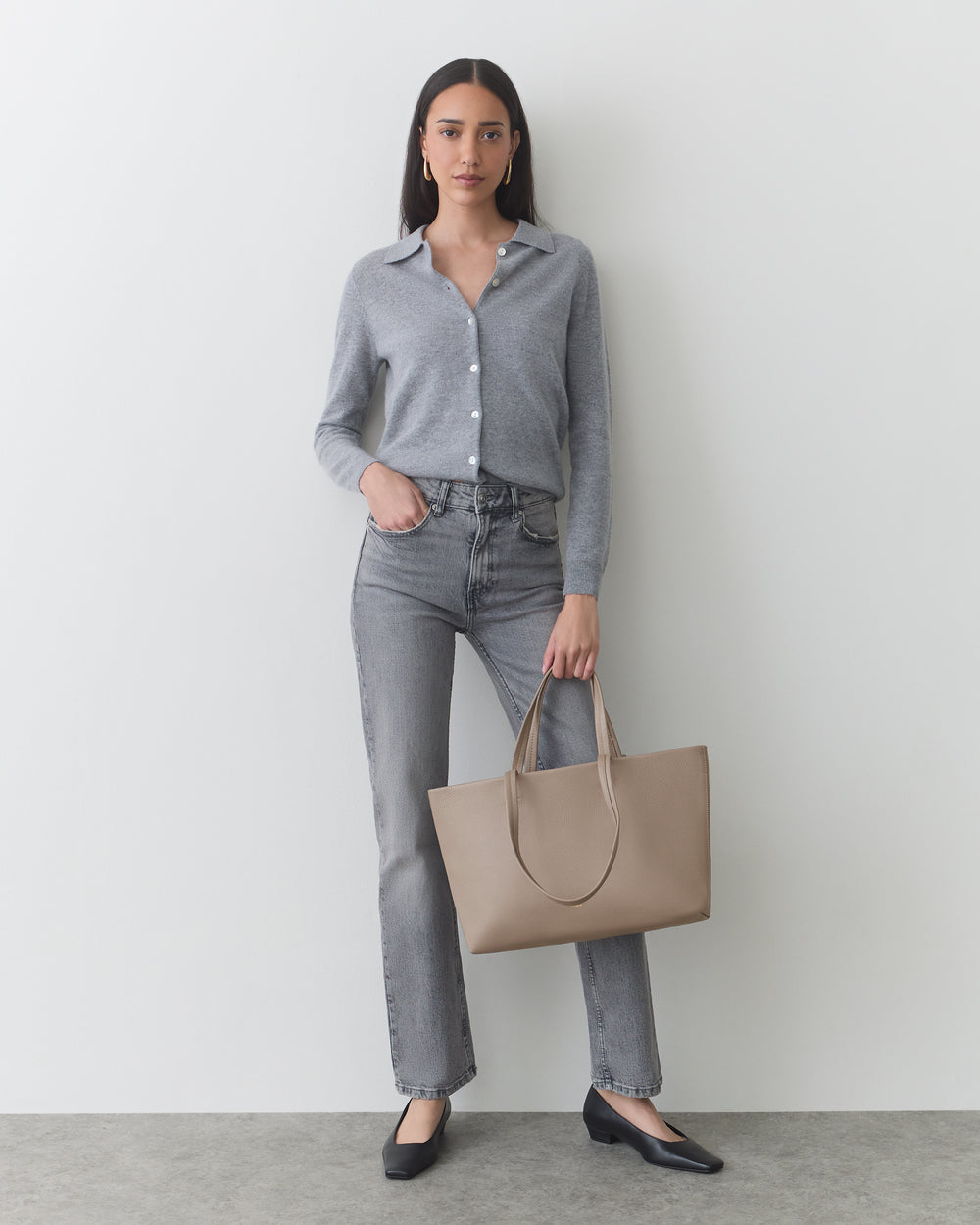 Person stands with neutral expression, wearing cardigan and jeans, holding a tote bag.