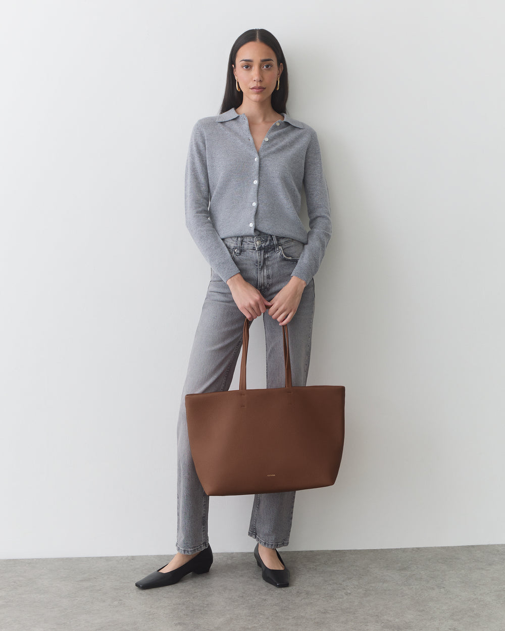 Person holding a large tote bag, wearing a button-up shirt and jeans, standing indoors.