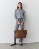 Person holding a large tote bag, wearing a button-up shirt and jeans, standing indoors.