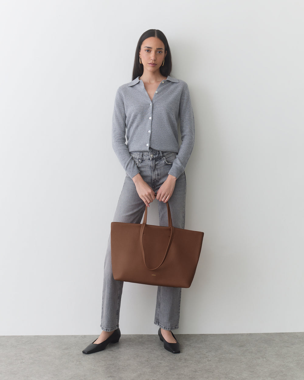Person standing, wearing cardigan and jeans, holding a large tote bag.