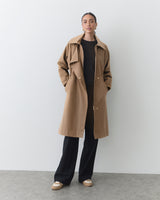 Person standing in long coat and pants, hands in pockets, against plain background.