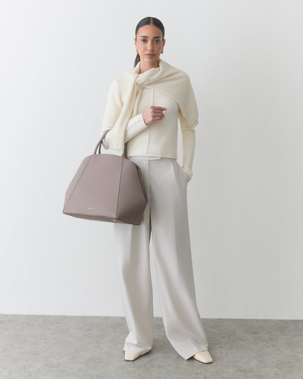 Person wearing a light outfit, holding a large handbag, standing against a plain background.