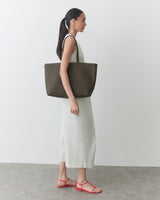 Person in a long dress carrying a large tote bag, standing on a plain surface.