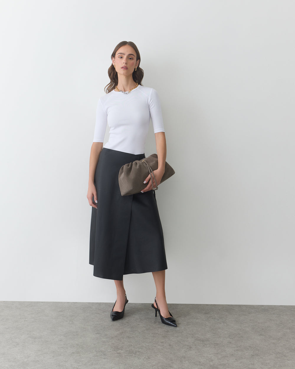 Woman in long skirt holding a curved pouch against a plain wall.