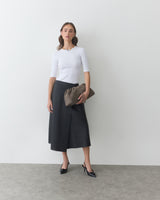 Woman in long skirt holding a curved pouch against a plain wall.