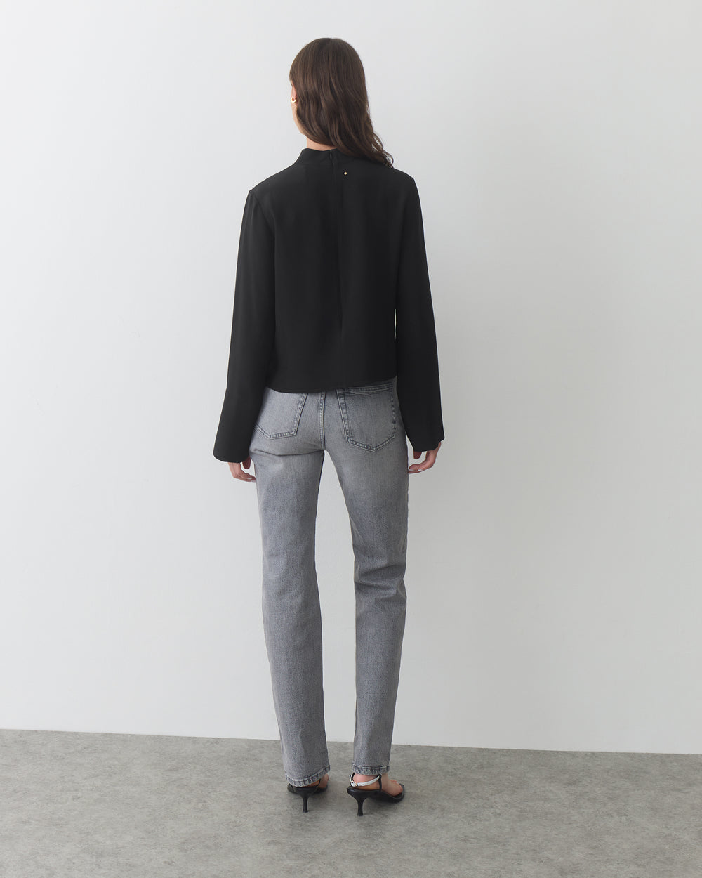 Person wearing long-sleeve top, jeans, and heels, standing in a plain indoor setting.