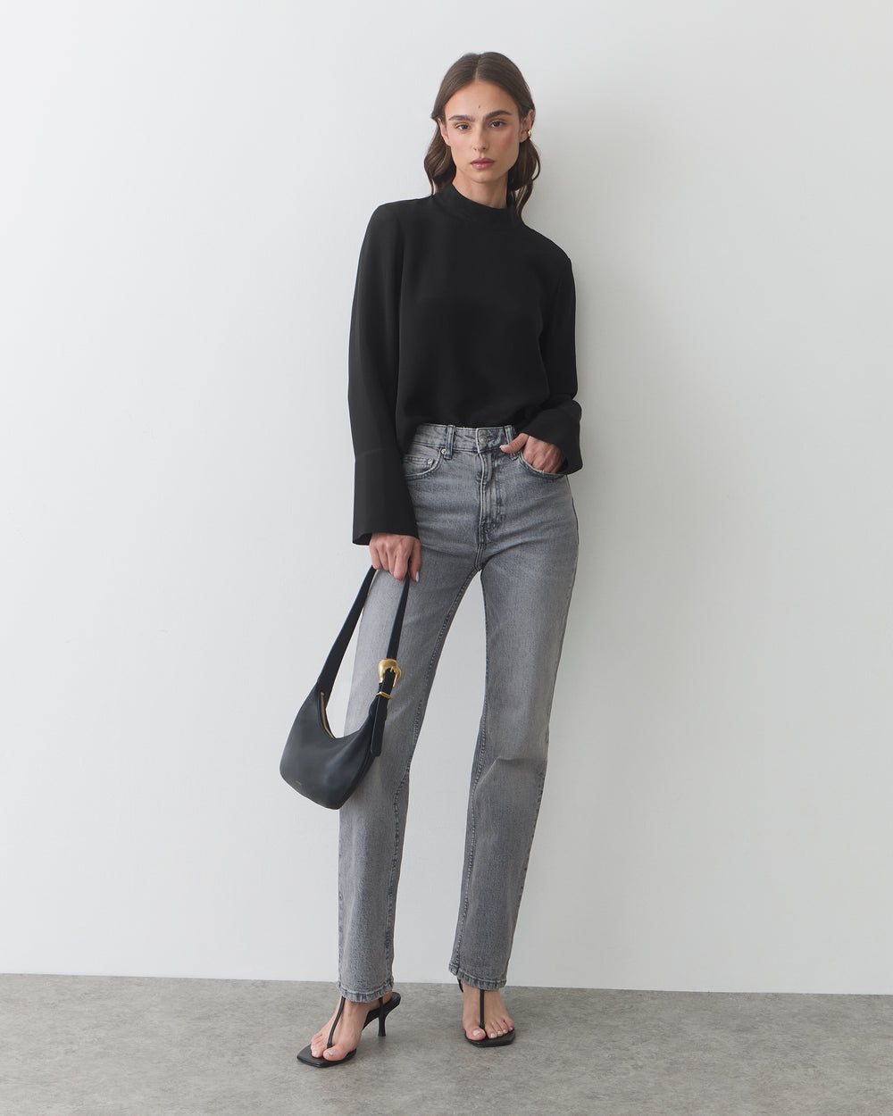 Person in long-sleeve top and jeans holding shoulder bag, wearing heeled sandals.