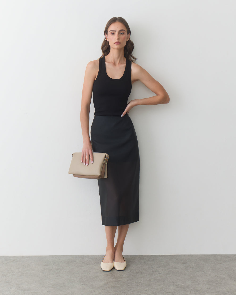 Woman in sleeveless top and skirt holds a handbag, leaning against a wall.