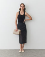 Woman in sleeveless top and skirt holds a handbag, leaning against a wall.